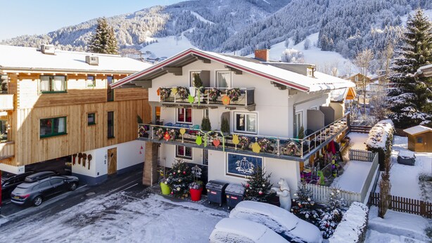 Un edificio di due piani con un balcone decorato con addobbi festivi, circondato da alberi e montagne innevate.