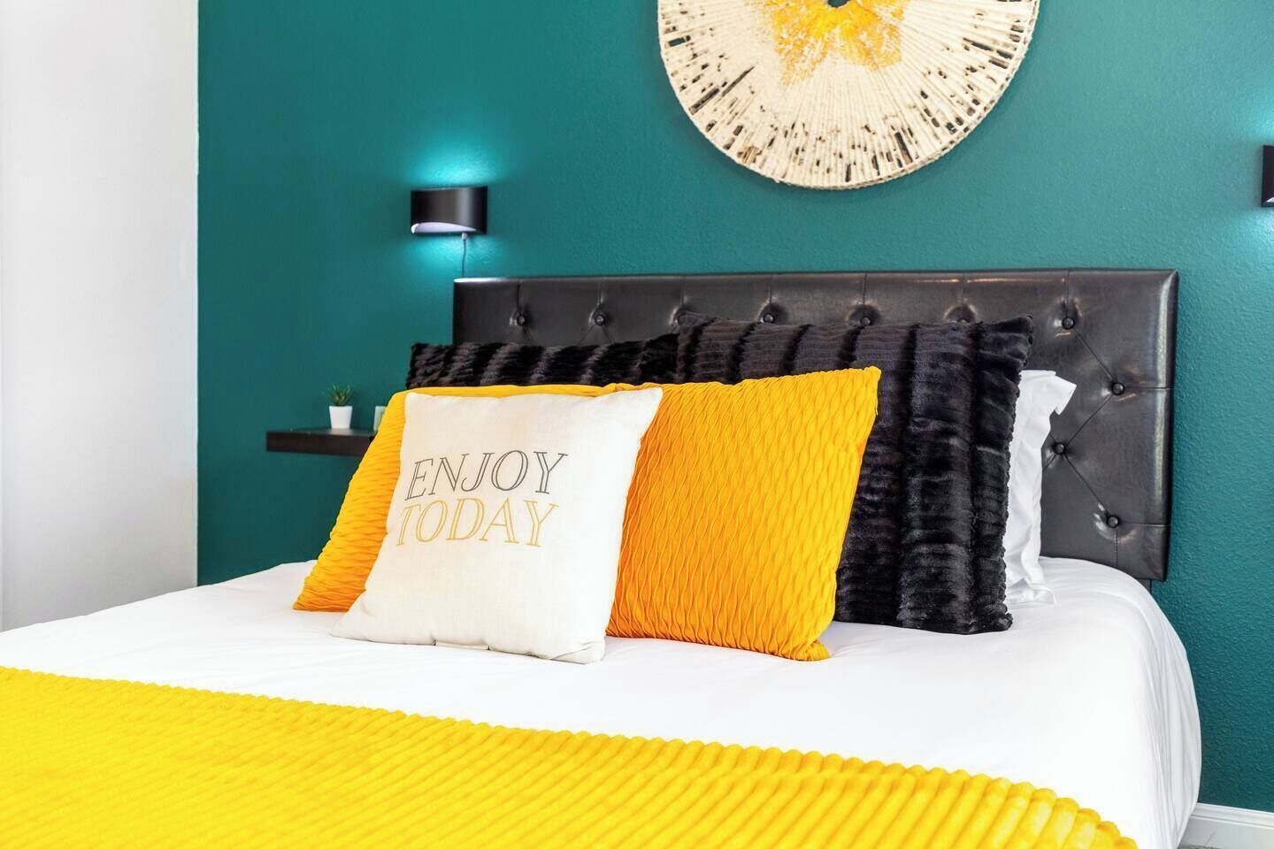 Un lit recouvert d’une couvre-lit blanche et jaune, deux oreillers ornés de motifs, une tête de lit sombre, une horloge murale et une petite plante posée sur une table de chevet.