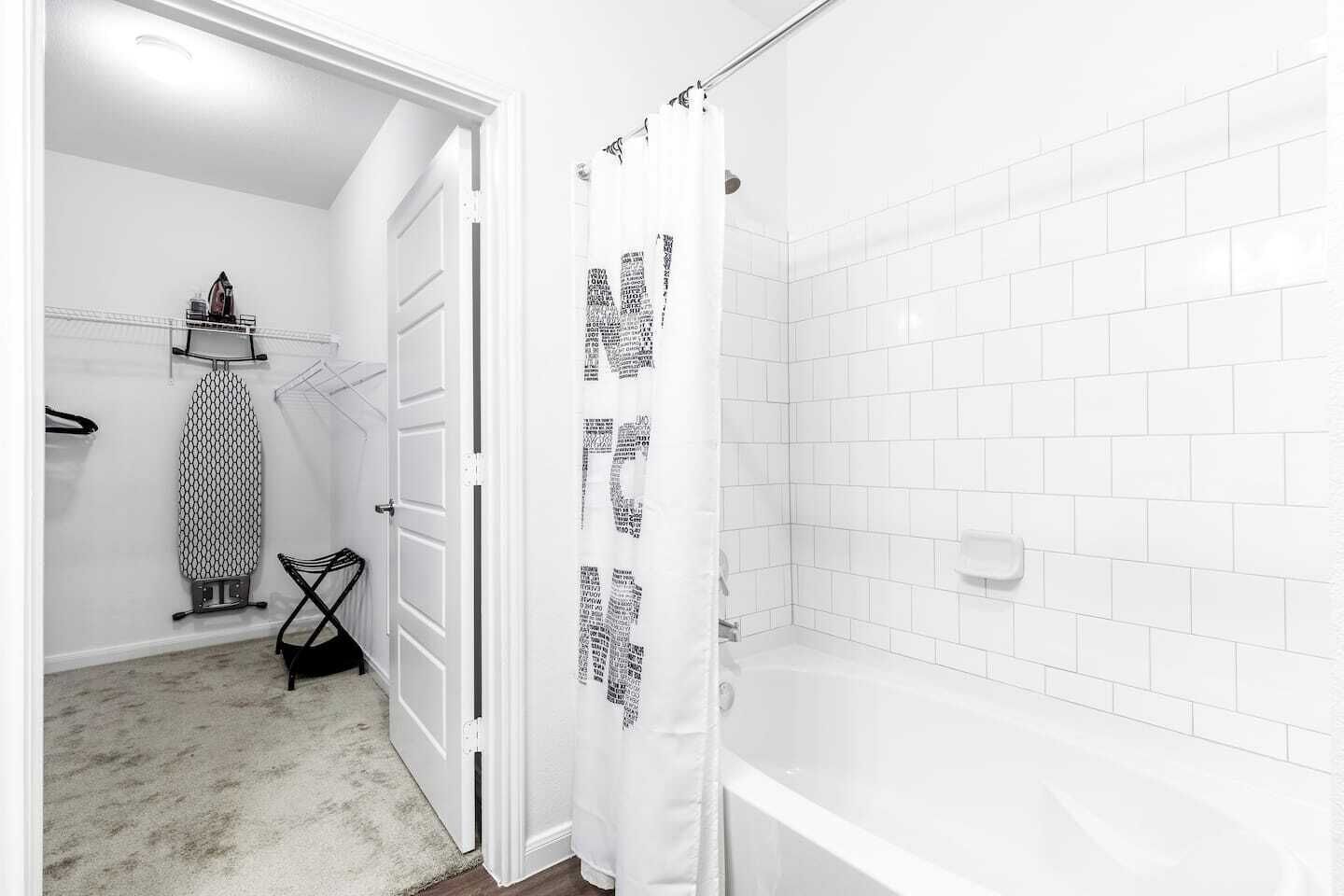 Une salle de bain avec des murs recouverts de carreaux blancs, un espace douche et une porte donnant sur une autre pièce équipée d’un portant à vêtements et d’une planche à repasser.