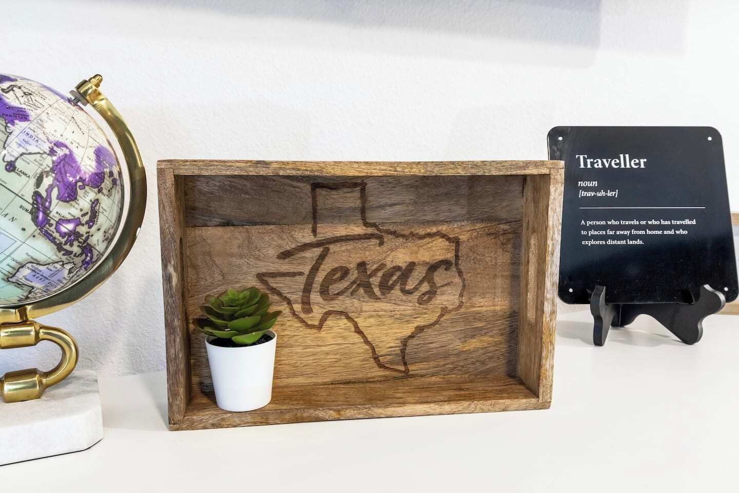 Une boîte en bois sur laquelle est gravé le mot « Texas », une petite plante en pot et une pancarte noire portant le mot « Traveller » et sa définition.