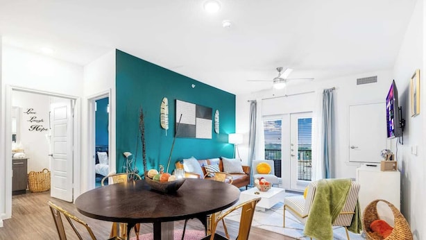 Ein modernes Wohnzimmer mit einer türkisfarbenen Akzentwand, einem Essbereich mit rundem Tisch und goldenen Stühlen sowie ein Balkon mit Aussicht.