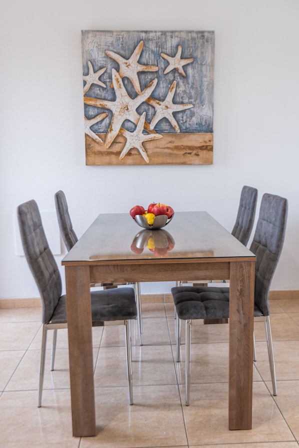 A dining table with a glass top, surrounded by four chairs. Above the table, there is a painting of starfish on a textured background. - Narcissos 'Nissi Beach' apartment C16 (Ayia Napa)