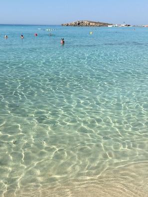 A clear beach with shallow water, a rocky outcrop in the distance, and boats on the water. - Narcissos 'Nissi Beach' apartment C16 (Ayia Napa)