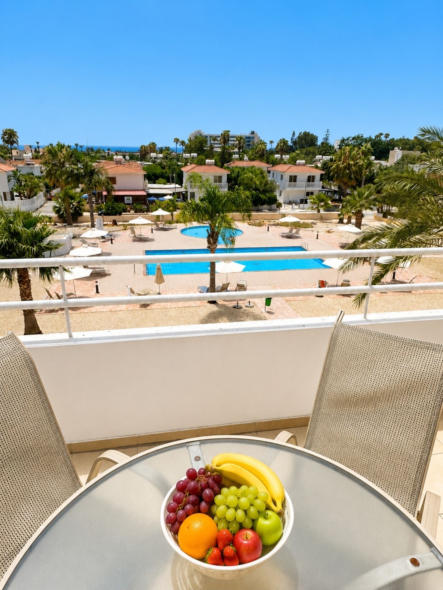 A balcony with a table holding a fruit bowl, overlooking a swimming pool and a residential area with palm trees and buildings.