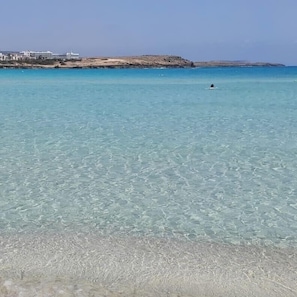 A clear coastal view with a sandy beach and a building in the background. - Narcissos 'Nissi Beach' apartment C16 (Ayia Napa)