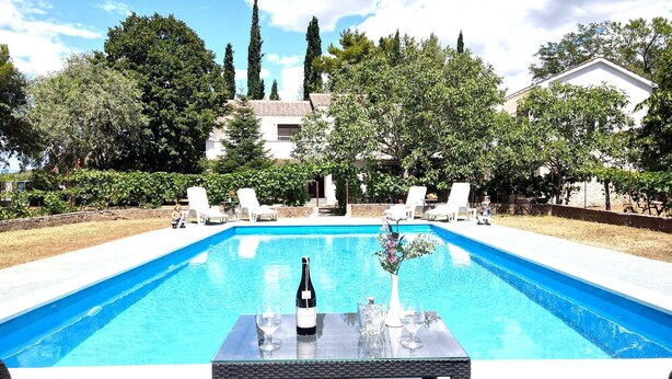 Une piscine rectangulaire à l’eau d’un bleu limpide, entourée de chaises longues blanches et d’une table sur laquelle se trouvent une bouteille de vin et des verres.