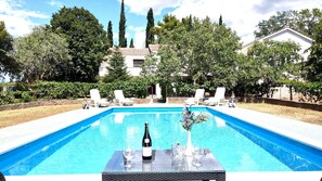 Una piscina rectangular con superficie de agua azul cristalina, rodeada de sillas de descanso blancas y una mesa con una botella de vino y copas.