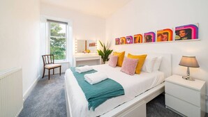 A bedroom with a bed, bedside table, chair, vanity with mirror, and wall art. - The Palais - Retro Style (Dundee)