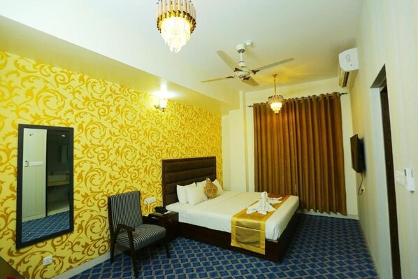 A hotel room with a patterned yellow wall, a large bed with a dark headboard, a chair, a ceiling fan, and a chandelier.