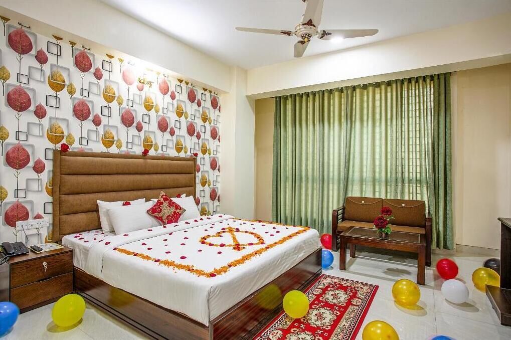 A hotel room with a bed, a wooden nightstand, a chair, a sofa, a ceiling fan, and a window with green curtains.