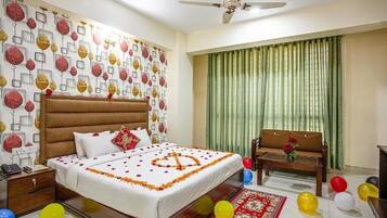 A hotel room with a bed, a wooden nightstand, a chair, a sofa, a ceiling fan, and a window with green curtains.