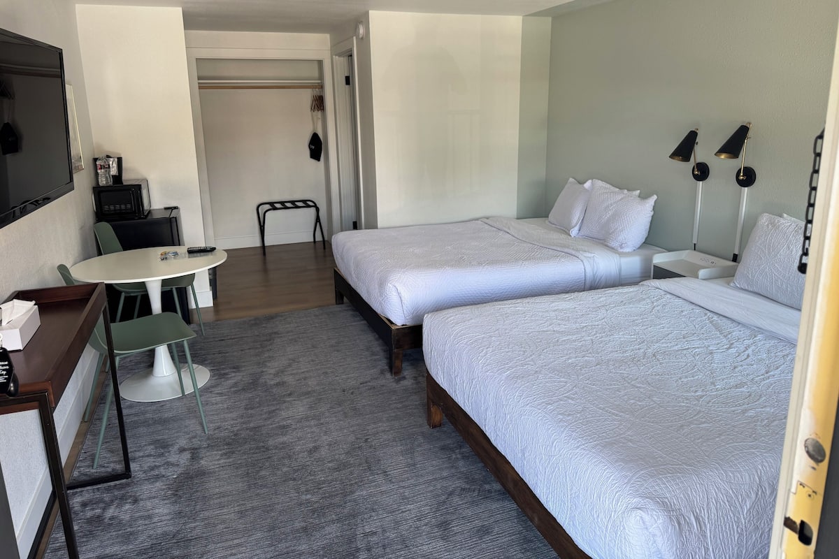 A hotel room with two beds, a desk with a chair, a small table, and a TV.