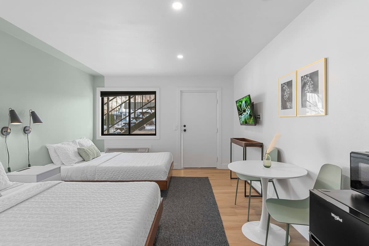 A modern hotel room with two beds, a desk, a chair, a TV, and a window with a view of a staircase.