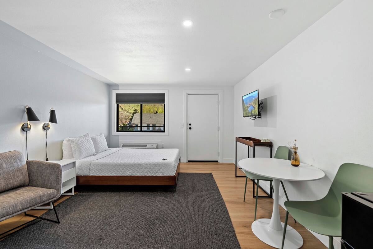 A modern bedroom with a bed, a sofa, a dining table with chairs, a TV, and a window with a view of trees.
