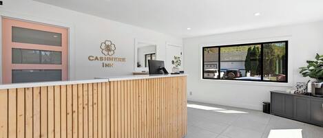 Et receptionområde med en træreception, et stort vindue med udsigt til en bygning og træer, samt et skilt der står 'KONTANTISTE'.