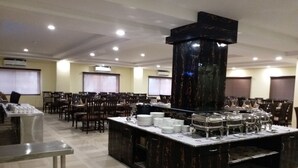 A spacious dining area with multiple tables and chairs, a central buffet station with various food items, and a large pillar structure. - The Sravasti Residency (Ikauna)
