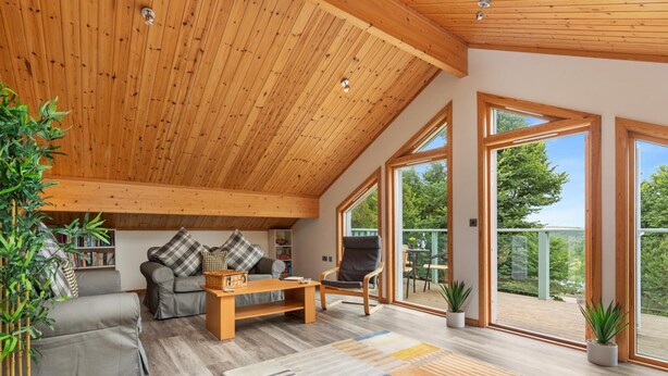 Un salon spacieux avec un plafond haut, des poutres apparentes, un canapé gris, une table basse en bois et de grandes fenêtres offrant une vue sur l’extérieur.