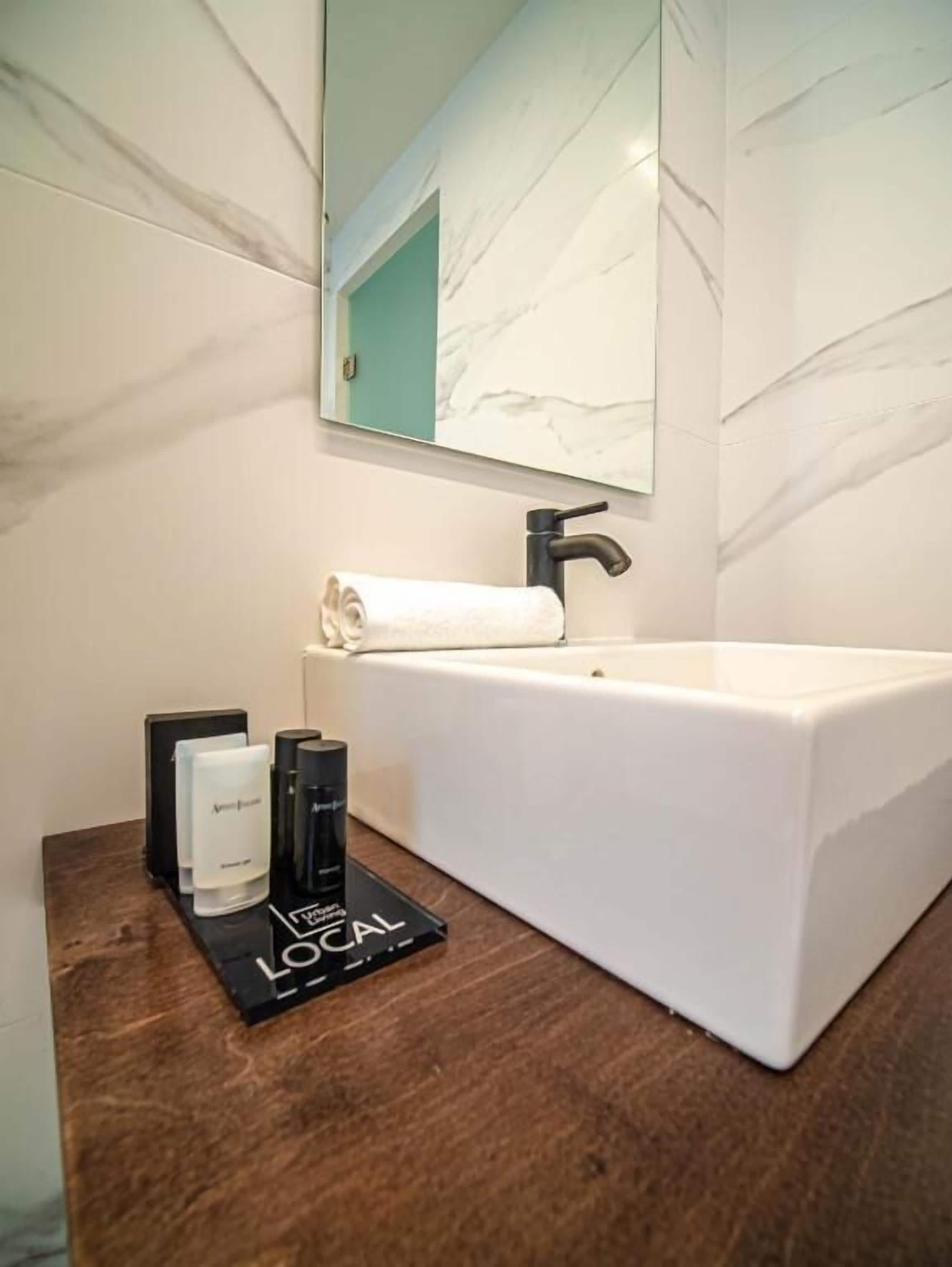 Une salle de bain avec un lavabo blanc, un miroir au-dessus et une serviette posée sur le comptoir. On y trouve également un distributeur de savon noir et blanc et une bouteille noire sur le comptoir.