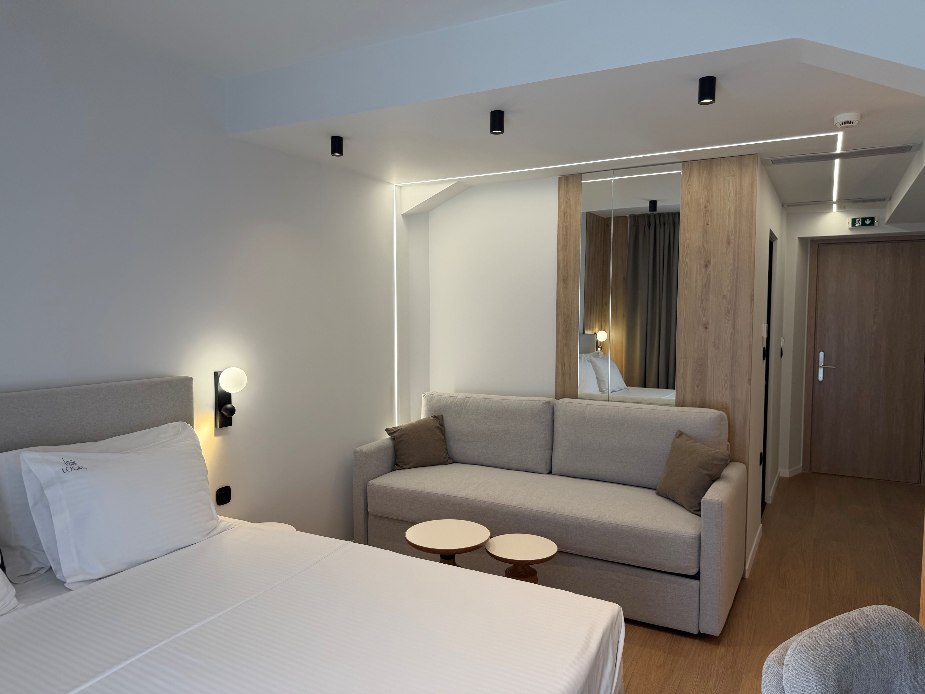 A hotel room with a bed, a sofa, two small round tables, a mirror, and a door.