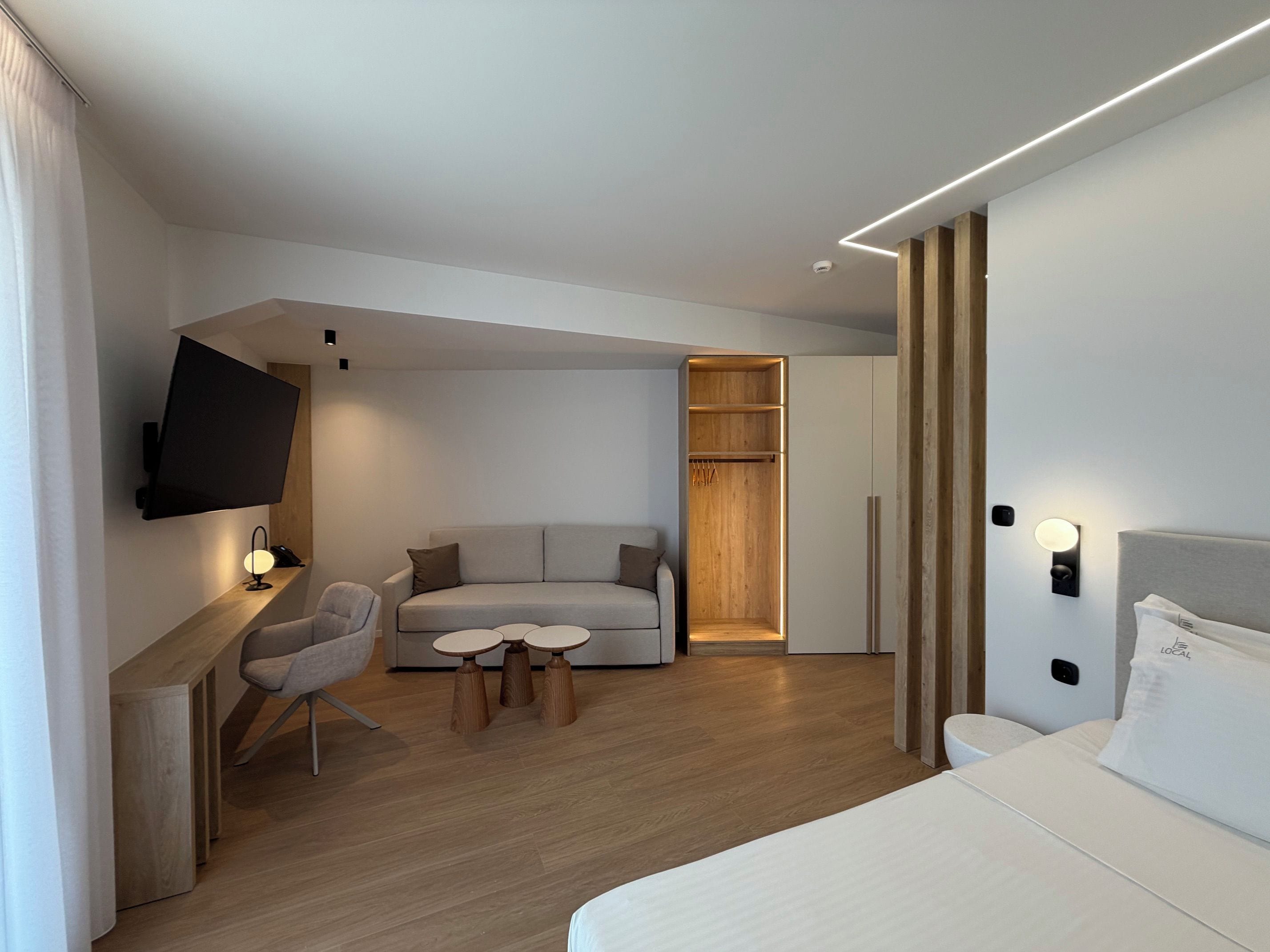 A modern hotel room with a flat-screen TV, a sofa, a dining table, a wooden wardrobe, and a bed.