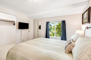 Um quarto com uma cama, televisão na parede, janela com cortinas e uma porta.