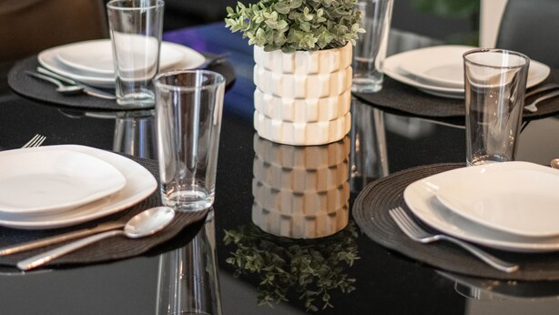 Ein Esszimmertisch ist gedeckt mit Tellern, Gläsern und Besteck, daneben eine dekorative Vase mit Grünpflanzen und ein an der Wand montierter Fernseher.