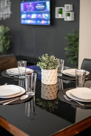 Une table à manger dressée avec des assiettes, des verres et des couverts, un vase décoratif orné de verdure, et un téléviseur fixé au mur.