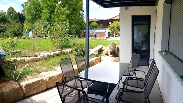 Un patio exterior con una mesa y sillas, un muro de piedra y vistas a un jardín y edificios al fondo.
