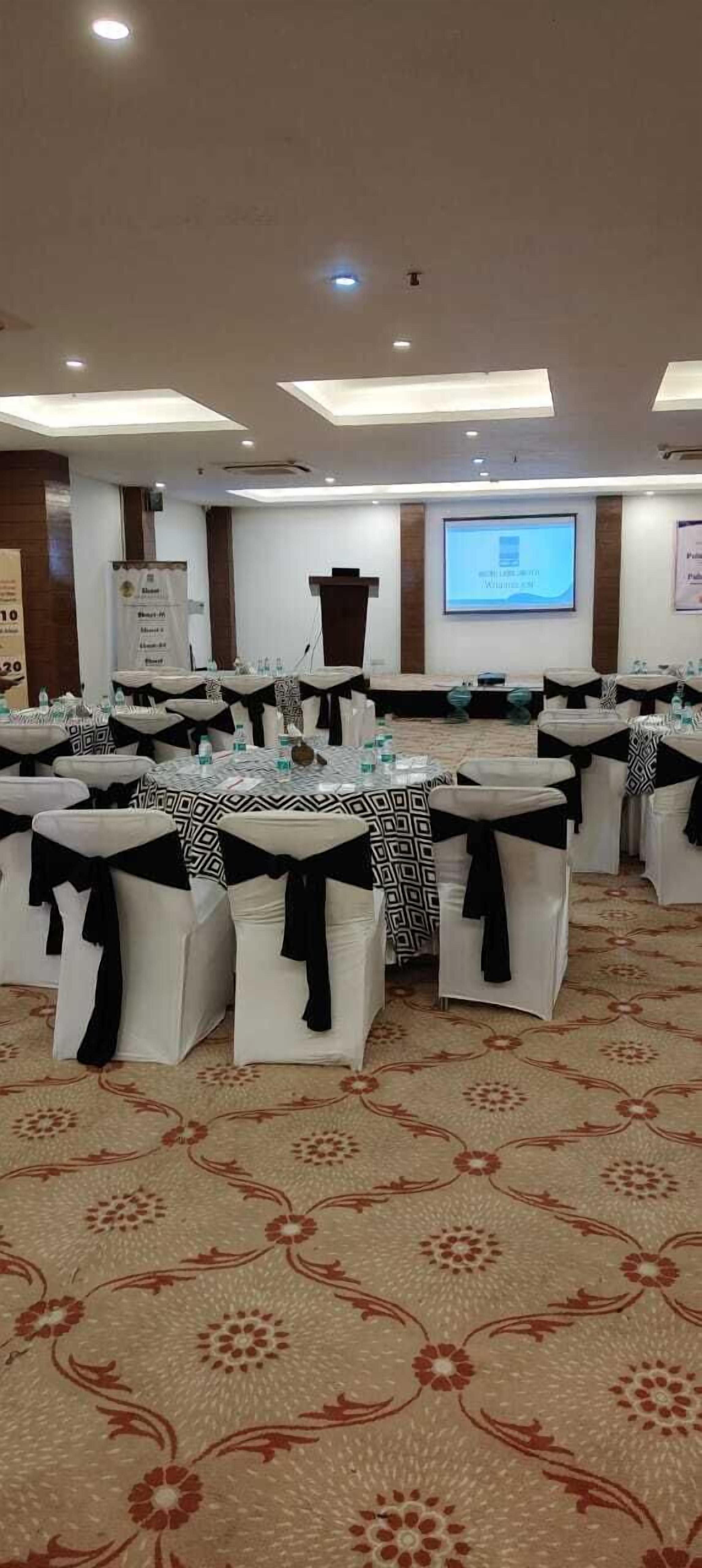 A conference room with round tables, each with a black and white patterned tablecloth and black sashes. The room has a carpeted floor and a projector screen on the wall.