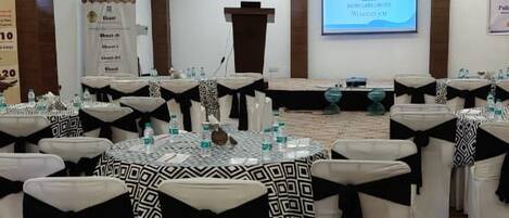A conference room with round tables, each with a black and white patterned tablecloth and black sashes. The room has a carpeted floor and a projector screen on the wall.