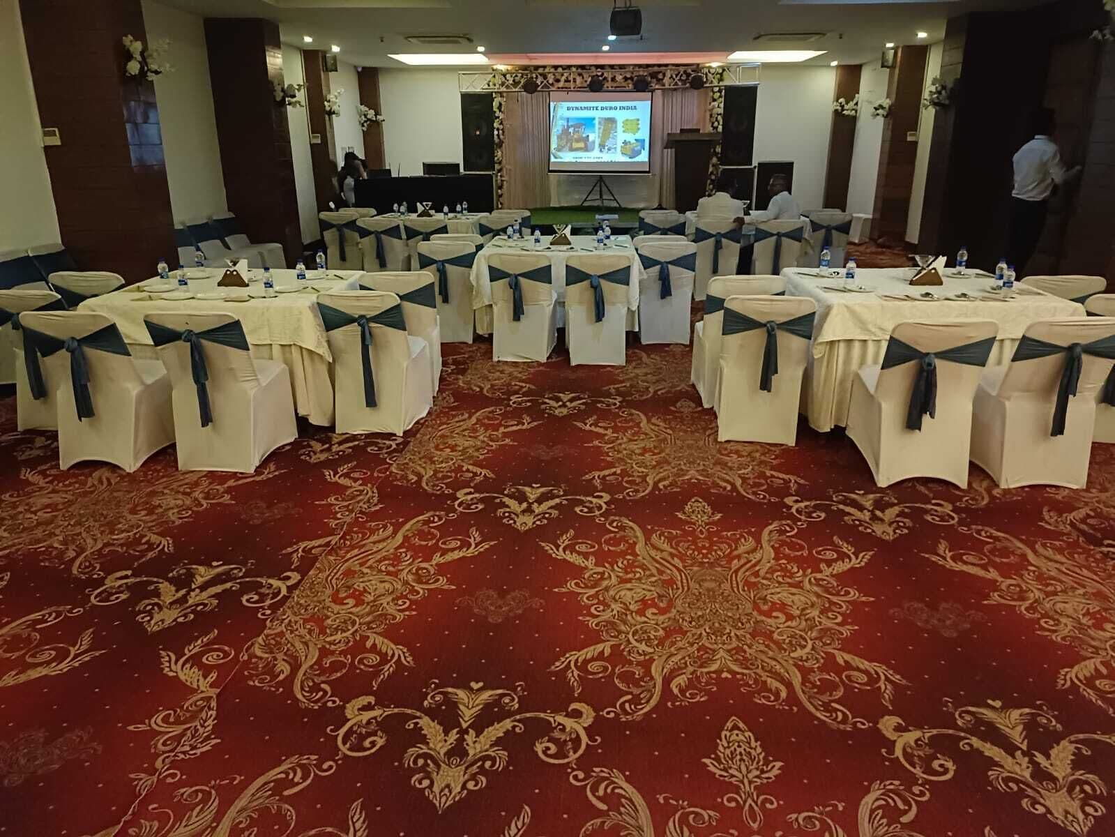 A banquet hall with round tables, chairs with blue sashes, a red carpet, and a projection screen in the background.