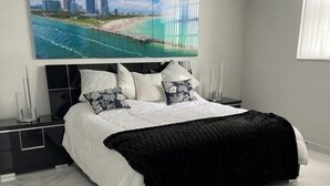 Ein modernes Schlafzimmer mit einem großen Bett, einer schwarzen Bank, einem Nachttisch und einem an der Wand befestigten Fernseher, der eine Strandansicht zeigt.
