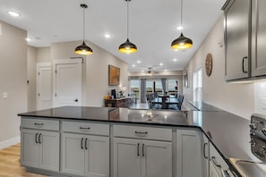 A kitchen with a central island, grey cabinets, stainless steel appliances, pendant lighting, and a dining area visible through an open door. - Ultimate Game Room Retreat w Putting Green_Great Lake Views_Free Tickets & Community In/Outdoor Pool (Branson)