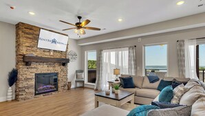 A spacious living room with a stone fireplace, a ceiling fan, and large windows offering a view of the outdoors.