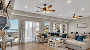 A spacious living room with a beige sofa, a coffee table, and a ceiling fan. There is a large window with curtains, a fireplace, and a view of a house.