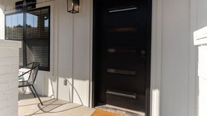 A black door with a metal handle, a window with blinds, and a chair outside.