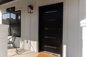 A black door with a metal handle, a window with blinds, and a chair outside.
