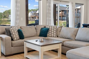 A modern living room with a sectional sofa, a coffee table, and a view of a residential area through large windows. - Near Silver Dollar City with Private Hot Tub, Game Area, Community Indoor Pool & Free Daily Tickets (Branson)