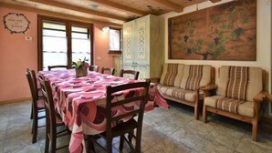 Un coin repas avec une longue table recouverte d’une nappe rose, entourée de chaises en bois. Un canapé avec des coussins à rayures est placé contre le mur, et un grand tableau représente un arbre avec des feuilles.