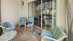 A covered porch with wicker chairs and a blue cushioned chair, a white door, and a large window with a view of the outdoors. - Tranquil Hideaway 4 Bdrm Condo in Avian Forest (Pawleys Island)