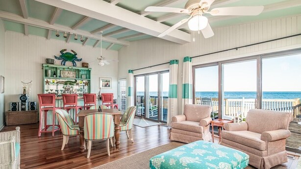 A spacious living room with a large window overlooking the ocean, a dining area with striped chairs, and a cozy seating area with a patterned ottoman.