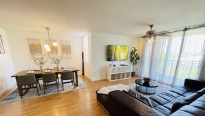 A modern living room with a black leather sofa, a dining area with a wooden table and chairs, a white entertainment unit, and a large window with sheer curtains.