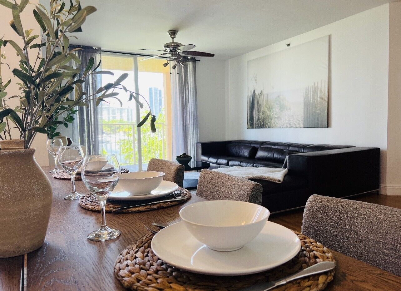 A dining area with a table set for two, a black leather sofa, and a ceiling fan.