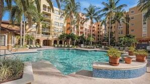 A hotel pool area with a clear pool, palm trees, and a multi-story building with balconies.