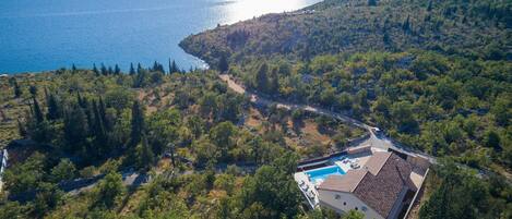 An aerial view of a property with a swimming pool, surrounded by dense forest and a body of water.