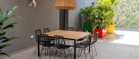 Un área de comedor al aire libre con una mesa de madera y sillas negras de alambre, rodeada de plantas en macetas y un colgador de luces.
