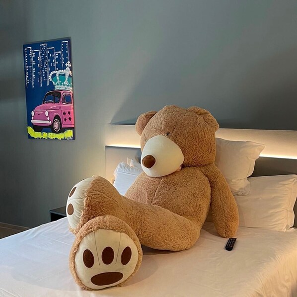 A bed with a large teddy bear and a smaller plush toy, a remote control, and a wall with a colorful painting.