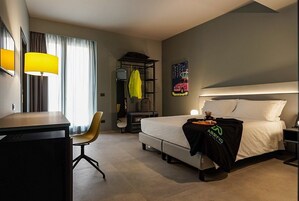 A hotel room with a bed, a desk, a chair, a wardrobe, and a poster on the wall.