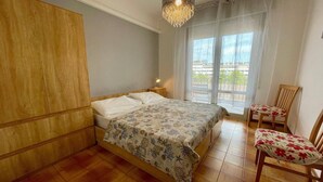 Ein Schlafzimmer mit Holzschrank, Bett mit floralem Überwurf, einer Chandelüster, einem Sessel mit Blumenkissen und einem Balkon mit Blick.