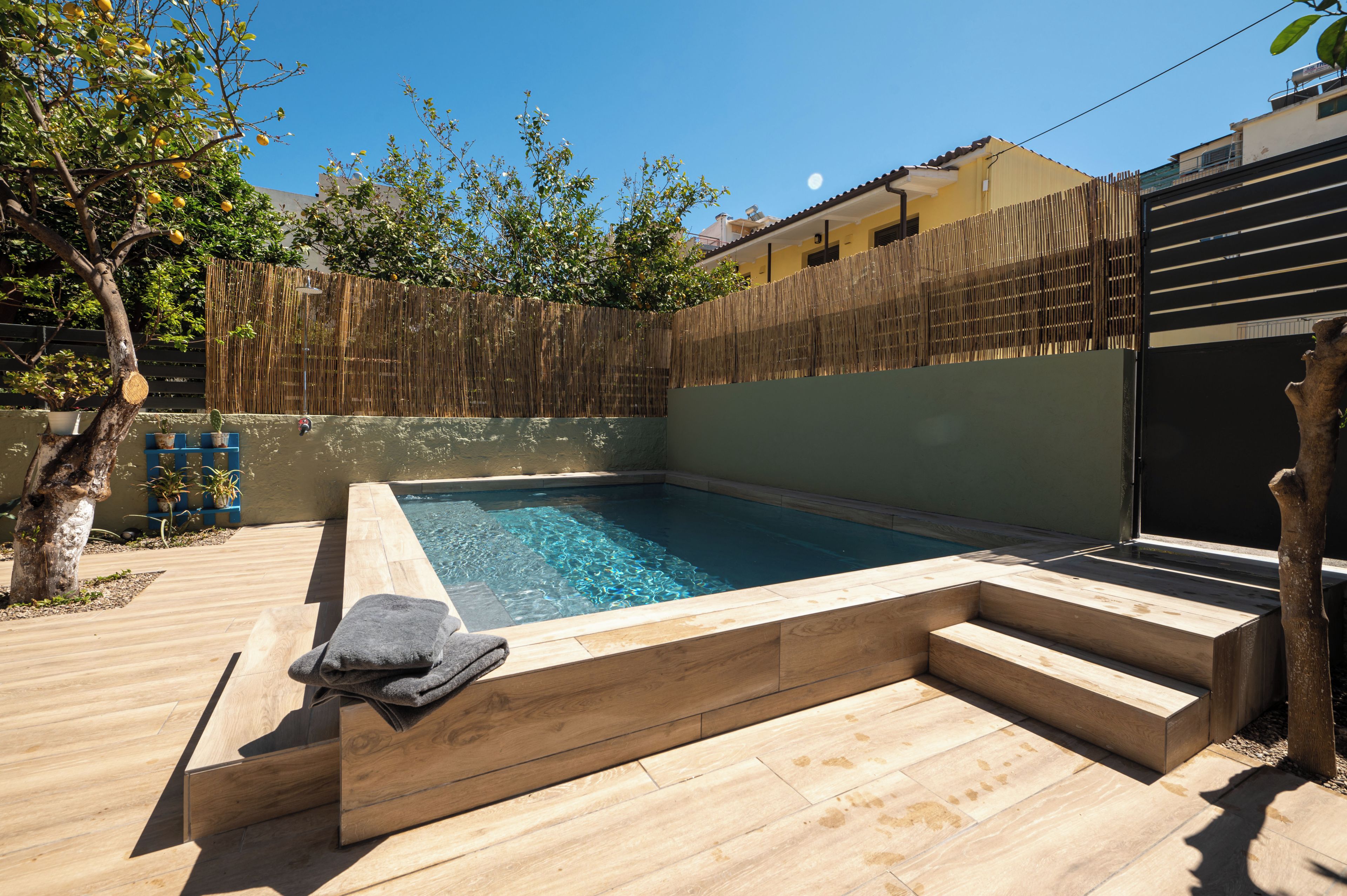 A rectangular swimming pool with a wooden deck and steps leading into it, surrounded by a bamboo fence and trees.